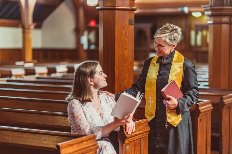 A female pastor converses with a congregant in a warmly lit church filled with wooden pews.