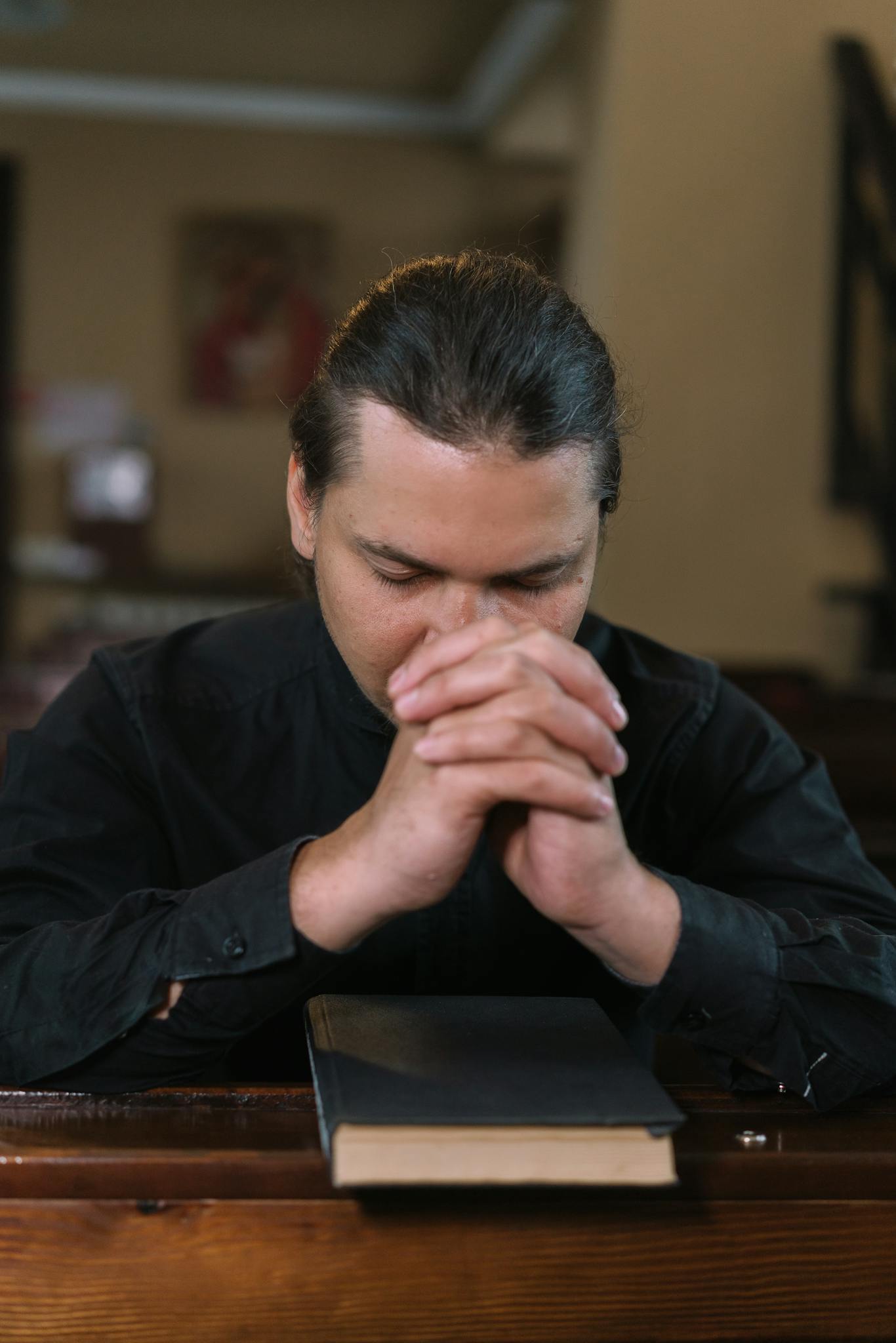 A man in black clothing prays in a church pew, hands interlocked with a holy book nearby.