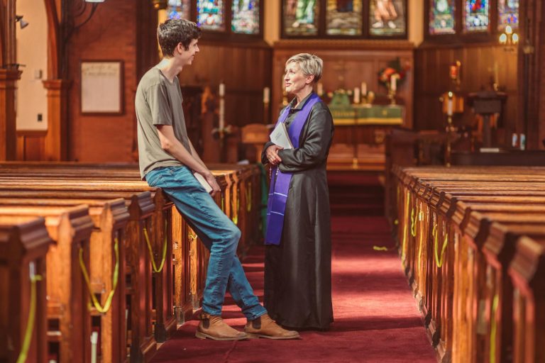 A priest converses with a young man inside a warmly lit church interior.