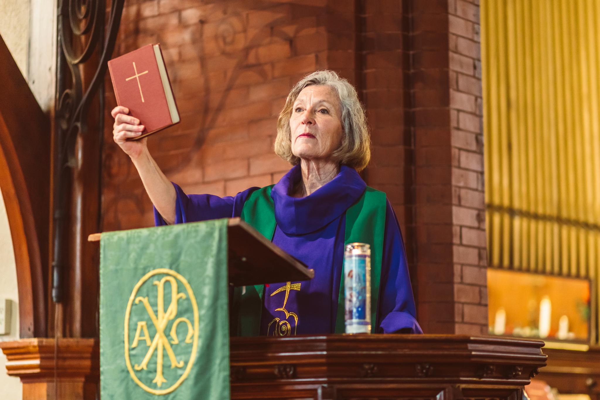 Elderly female priest delivering a sermon in a traditional church setting, holding a Bible.