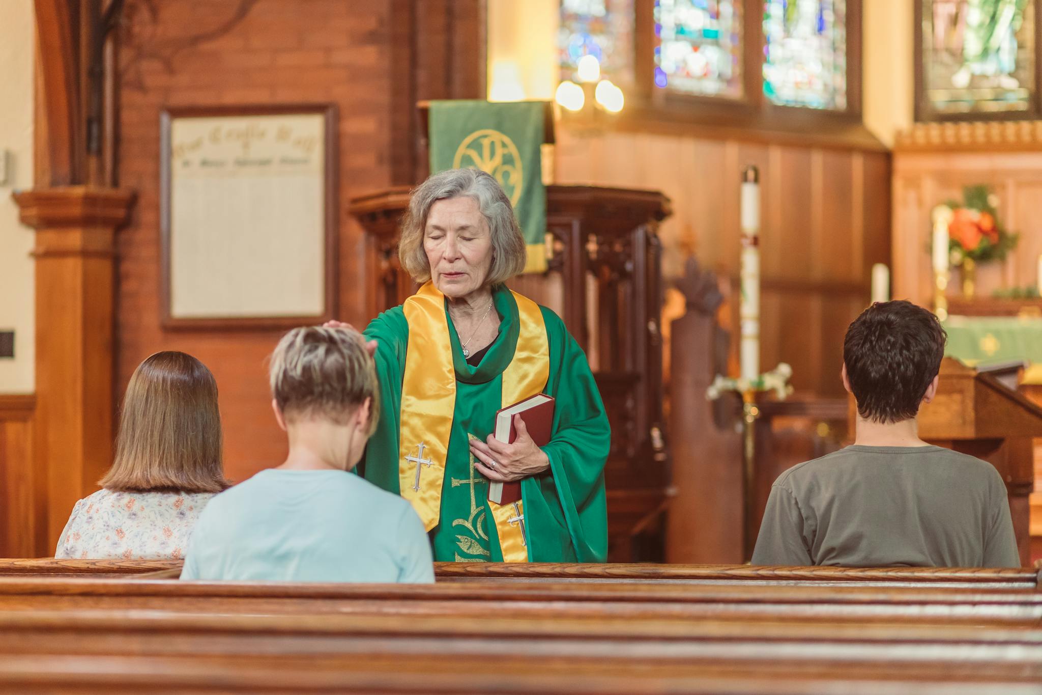 Elderly woman in clergy robe leads prayer in a church setting, surrounded by worshippers.