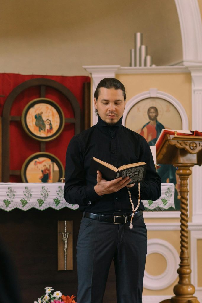 Priest in black attire reading a bible with prayer beads during church ceremony.